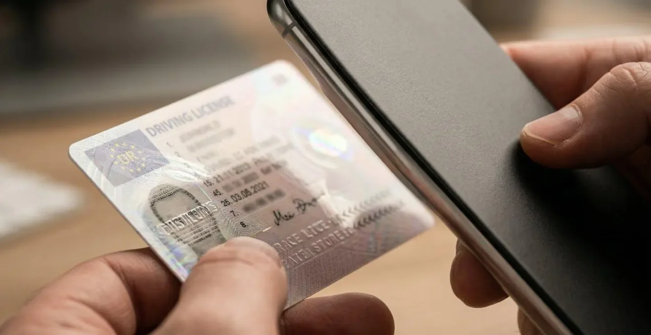 Main tenant un smartphone montrant une application floue à l'arrière-plan, avec un permis de conduire physique au premier plan